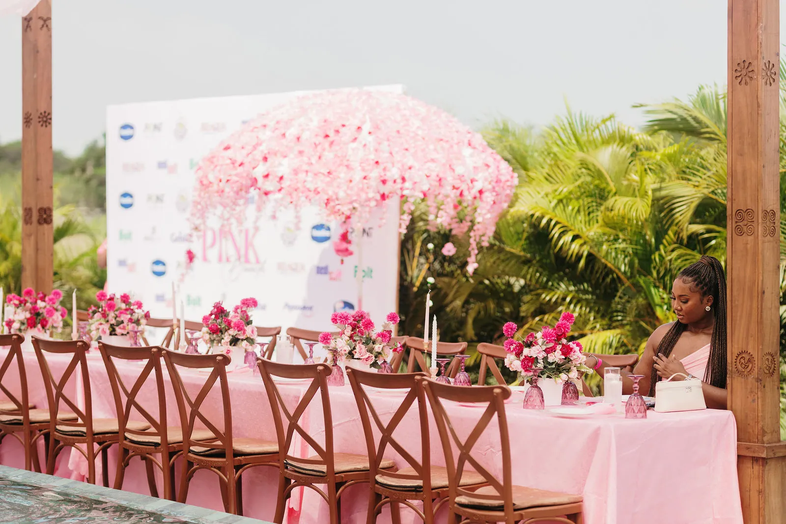 Long brunch table dressed in pink linen and flowers beneath a pink floral canopy, with palm trees behind